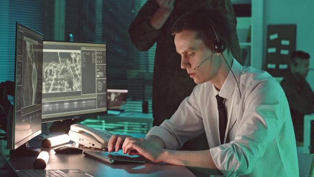 Young male security office in microphone headset tracking target while watching footage from satellite on computer display in dark central office hub