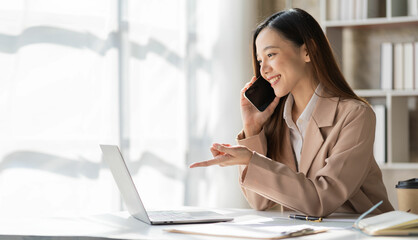 Attractive smiling asian businesswoman in light brown shirt talking on the phone with customer to...