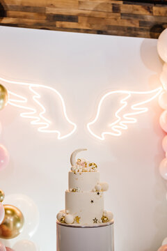Trendy cake with decor. Celebration baptism concept. Arch decorated with pink, white, golden balloons, angel wings. Reception at birthday baby party on wall. Delicious reception on photo zone, area.