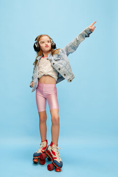 Music. Little beautiful girl, child in shorts and jeans jacket posing in headphones on rollers over blue studio background. Concept of childhood, emotions, fun, fashion, lifestyle, facial expression