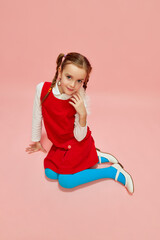 Cute, beautiful little girl, child with two pigtails posing in red dress and blue tights over pink studio background. Concept of childhood, emotions, fun, fashion, lifestyle, facial expression