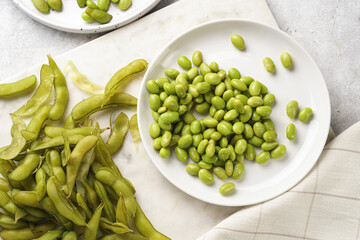 Peeling fresh steamed green soy beans edamame. Japanese plant snack on marble board on light grey background