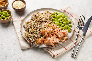 A flat grey plate with quinoa tricolore, roughly chopped and pulled salmon and steamed green soy beans edamame with sesame seeds and deep fried onions on light background. Top view