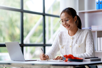 Beautiful Asian businesswoman using her laptop to work and enjoy taking notes on meeting minutes agendas of secretarial work.