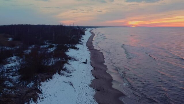 Static aerial view of a lonely man standing in snowy daugavgriva beach while waves breaks smoothly in the shore at sunset