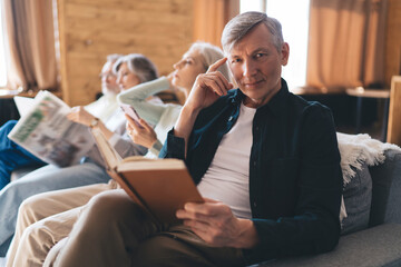 Positive senior man resting on couch with book spending time with friends
