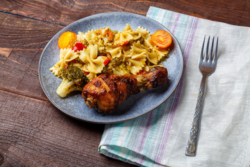 Pasta with vegetables and cherry tomatoes with baked chicken leg on the table next to a light napkin and fork.