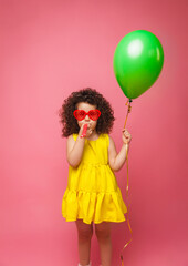 Happy birthday celebration with flying balloons of a charming cute little girl in a yellow dress isolated on a pink background.
