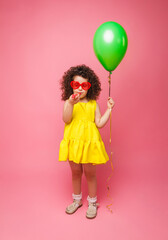 Happy birthday celebration with flying balloons of a charming cute little girl in a yellow dress isolated on a pink background.