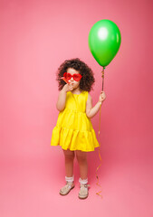 Happy birthday celebration with flying balloons of a charming cute little girl in a yellow dress isolated on a pink background.