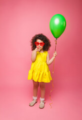 Happy birthday celebration with flying balloons of a charming cute little girl in a yellow dress isolated on a pink background.