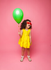 Happy birthday celebration with flying balloons of a charming cute little girl in a yellow dress isolated on a pink background.