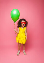 Happy birthday celebration with flying balloons of a charming cute little girl in a yellow dress isolated on a pink background.