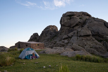 Camping in the top of the rock massif Los Gigantes in Cordoba, Argentina. View of a young woman, tent, Nores refuge and the rocky hills at sunset. 