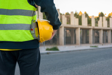Unrecognizable worker with yellow hard hat in hand observes a building under construction