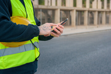 Close-up of a laborer looking at a phone while holding a yellow hard hat with a construction site in the background