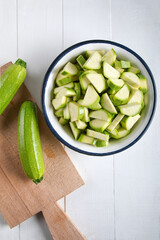 Two green zucchini on a cutting board with chopped zucchini in a bowl on a wooden background.