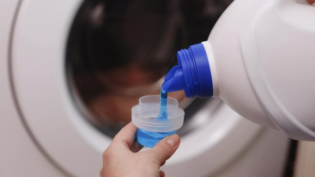 Close up shot of a woman hands squeezing liquid gel laundry detergent from a plastic bottle into a small container, with a washing machine in the background. 4k Slow motion.