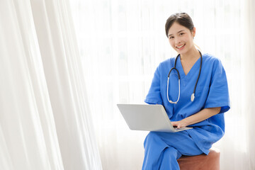 Portrait of a young Asian happy nurse wearing medical scrubs looking at the camera with a stethoscope and using a laptop while sitting with a white curtain in the background. Image with copy space.