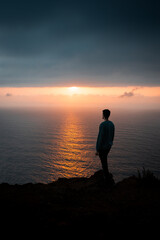 a view from a drone of the ocean and a man standing and looking into the distance, and the sun hidden in the clouds