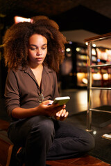 portrait young afro black brazilian woman sitting on a step holding and reading her cell phone in a library cafeteria