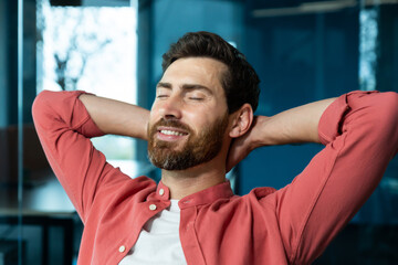 Successful businessman in office in a red shirt rests with his hands folded head with closed eyes,...