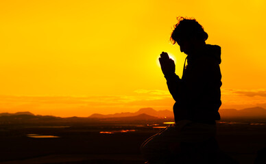 Silhouette of a male praying on top of a mountain at sunset with the sun just behind his hands. Concept of spiritualism and religion. Copy space on the right