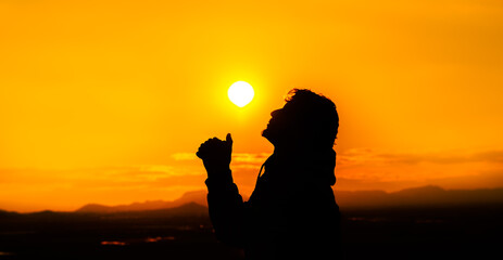 Silhouette of a person with joined hands, looking at the sky and praying. The sun is above him being the light that illuminates him. Concept of religion and prayer. Warm atmosphere at sunset.