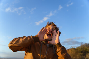 Fototapeta premium Young man shouting energetically to the sky with his hands over his mouth simulating a megaphone to project his voice. Front view. Copy space to the left.