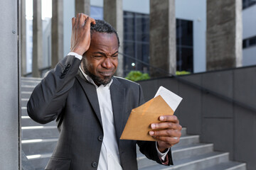 Upset African American man standing near the courthouse, holding an envelope with a letter in his hands. He holds his head, Received documents for divorce, financial agreement, alimony, lawsuit.