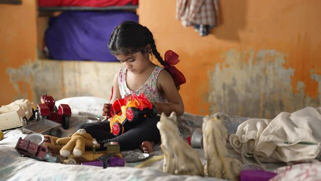 Preschooler or a young orphan child with long tail hair on bed look sad play with plastic toy car. Indian lonely little pretty unhappy village girl kid scolded spend leisure time activity at home