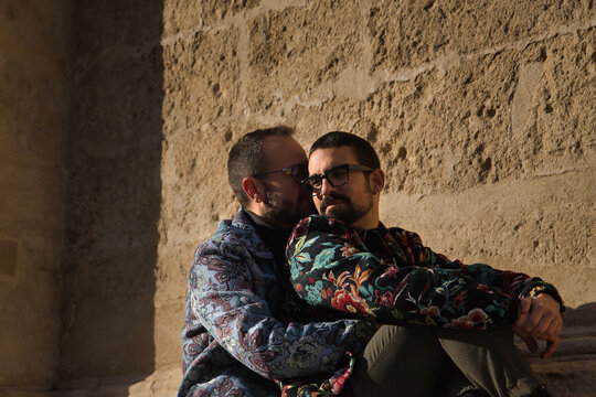 Real marriage of gay couple, sitting on a stone wall while one embraces the other from behind with very affectionate, complicit and happy attitude. Concept lgtb, lgtbiq+, couples, in love, love.