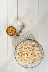 bowl with popcorn, popcorn kernels and salt on a checkered napkin , on a white wooden table, top view, copy space