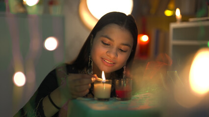 Smiling pretty Indian girl lighting colorful glass candles with matchstick on the occasion of...