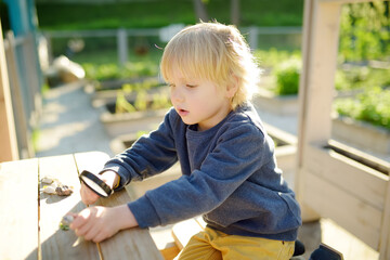 Child playing with pebbles and explores them with a magnifying glass. Little boy studying of various natural materials. Kids explore nature. Alternative education. Montessori educational system.