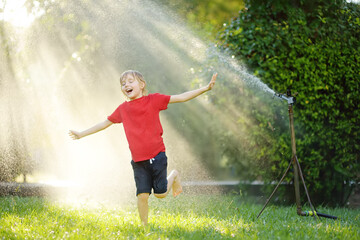 Funny little boy playing with garden sprinkler in sunny backyard. Elementary school child laughing,...