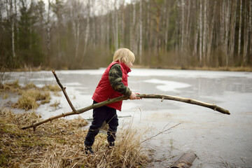 Little boy playing large branch on shore of forest lake on early spring day. Surface of lake is...