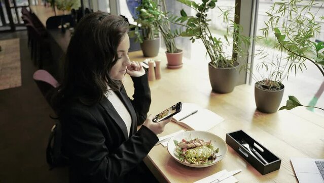 Young Caucasian Beautiful Woman Blogger Sitting In Restaurant At A Wooden Table In Front Of Huge Window. Girl Is Eating, Enjoying Her Meal, And Taking Pictures Of Her Food.