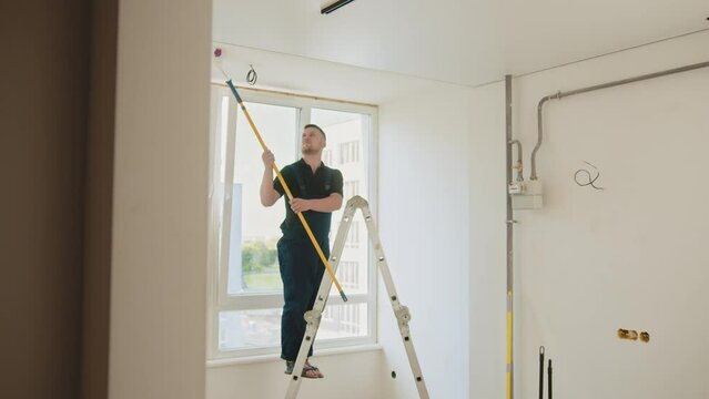 A bearded construction worker in a blue overall paints the ceiling in an apartment. The artist is a person. He uses a roller. A man paints the walls and ceiling in a new apartment. Repair.