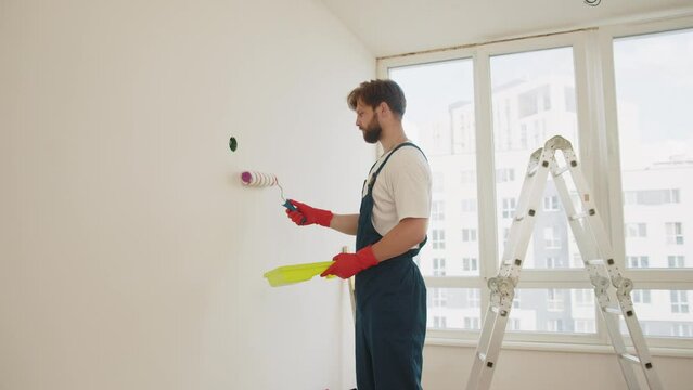 Close up of male in gloves holding painting roller. Painter man painting the wall in home, with paint roller and white color paint. Room renovations at house. Young worker painting wall in room.