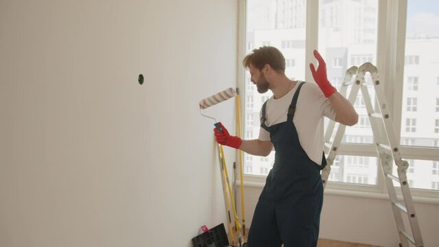 Young man, who have finally collected the necessary amount of funds for repairs, rejoice and dance. Instead of real microphone, guy using into a mop and a paint roller. House renovation concept.