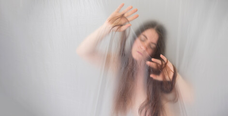 Fototapeta premium banner blurred, fuzzy image of sensual romantic young naked woman portrait behind plastic film, looking down, playing shy with her hair, moves in ecstasy