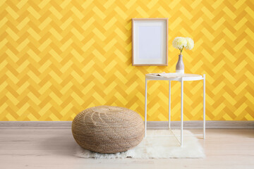Table with flowers in vase and pouf near yellow wall in room
