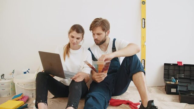 Happy Couple Choosing Design Of Their New Flat, Using Color Palette And Laptop While Sitting On Floor, Spouses Planning House Decoration Together. Planning new interio. Idea for repair