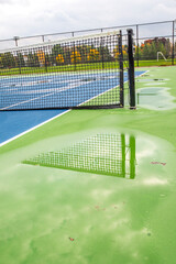 A tennis net is reflected in a pool of rain water on a blue and green colored court.