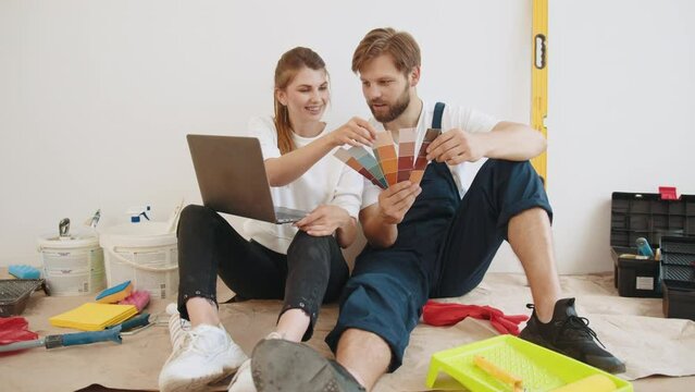 Joyful young wife and husband family couple sit on floor in new apartment during renovation process choosing new wall color and surfing internet on laptop choosing room design. Planning new interio