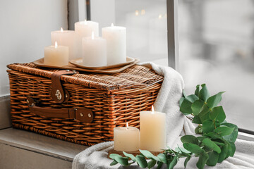 Burning candles, wicker case, green branch and woollen cover at windowsill