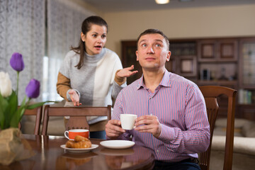 Adult man drinking tea at home table and ignoring disgruntled wife standing behind and reprimanding him