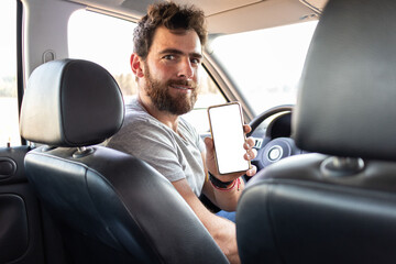 young man smiling showing the screen of his phone, sitting on driver's seat, app driver user's pov