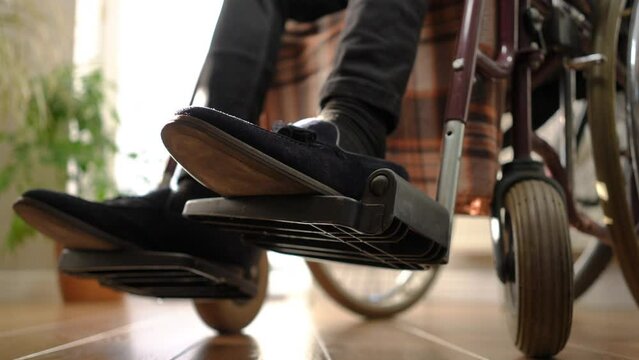 Close-up leg of young man in wheelchair on leg rest with female hand putting on shoe leaving. Unrecognizable woman taking care of spouse at home indoors. Rehabilitation and love concept